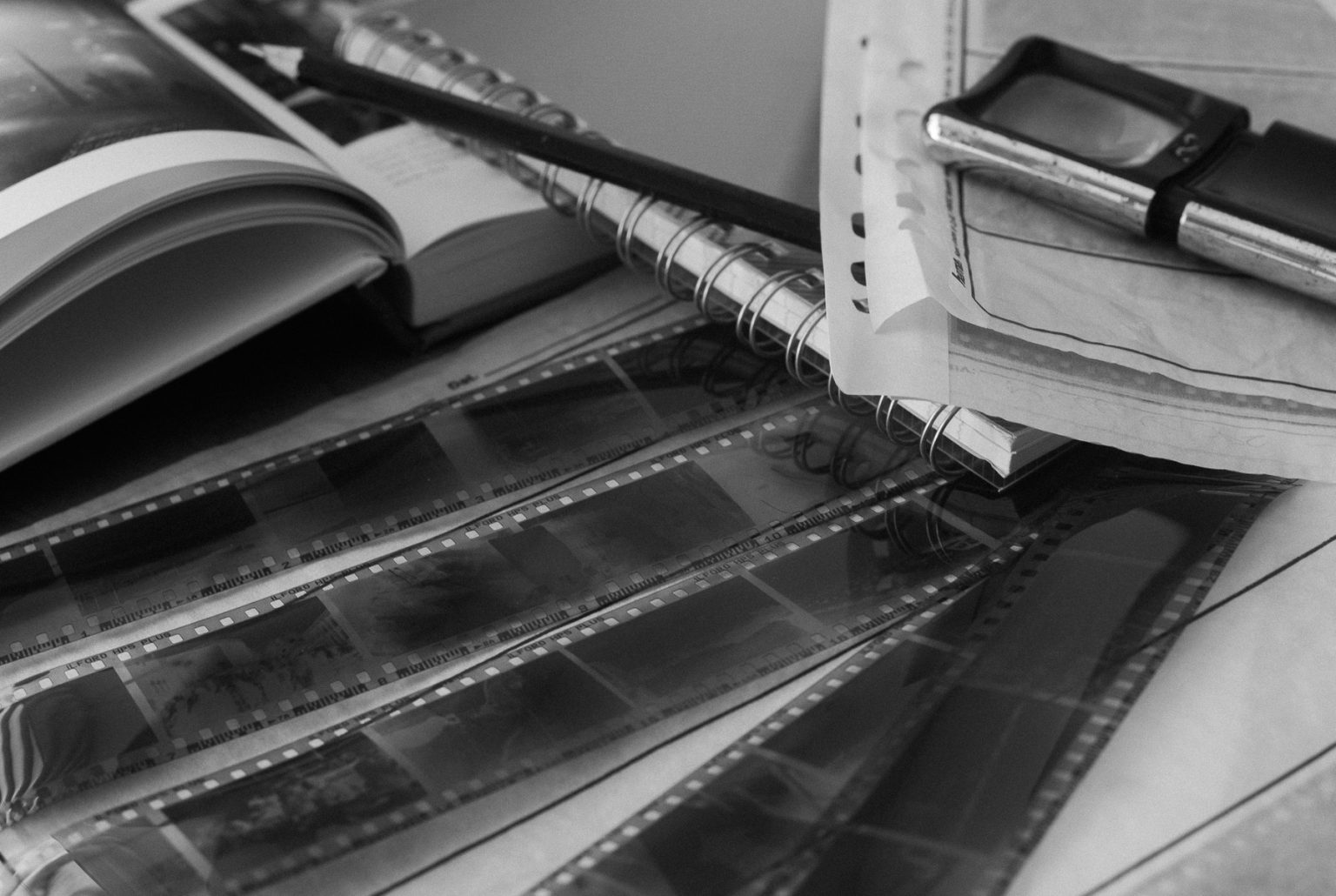 A black and white photo of film negatives, a loupe, a pencil, and a notebook on a wooden desk — a still life about photography’s relationship with time and memory.